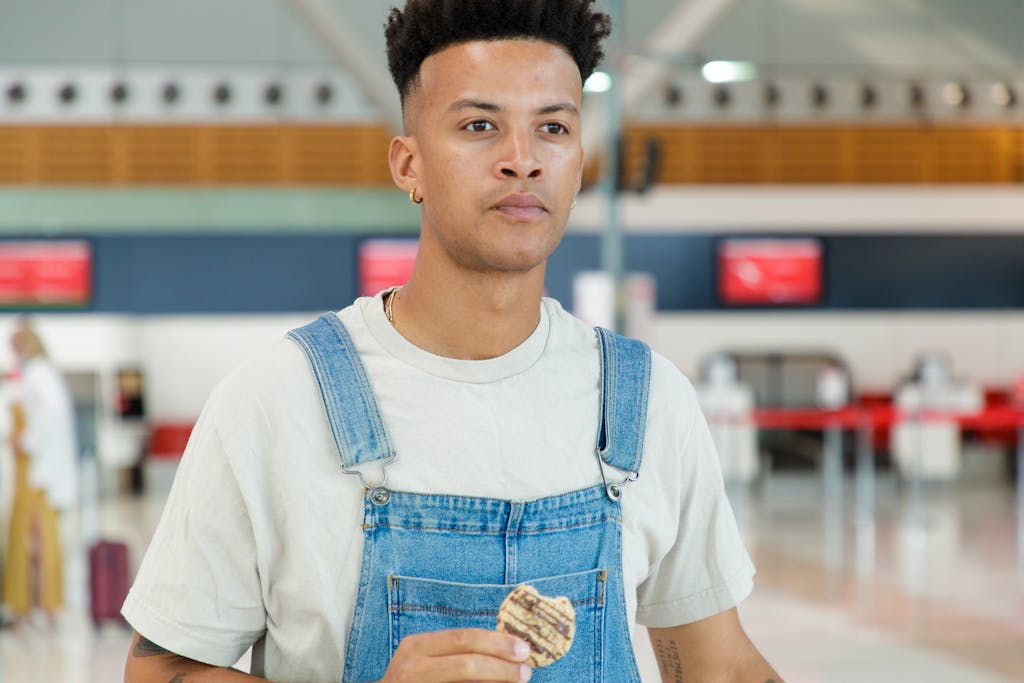 Casual young man in denim overalls eating a cookie at an airport terminal.