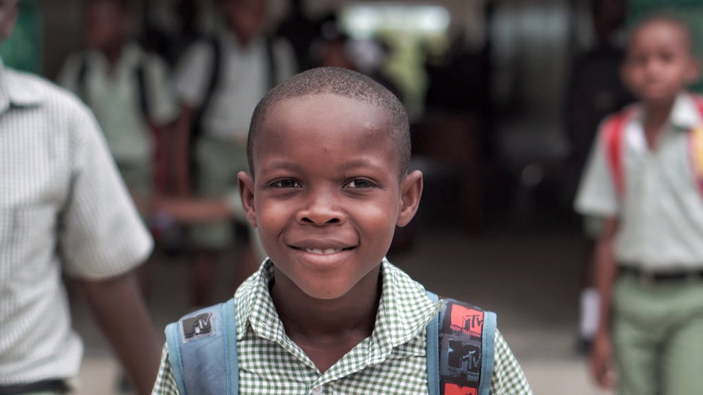 A cheerful schoolboy in a Haitian school uniform, carrying a backpack outdoors.