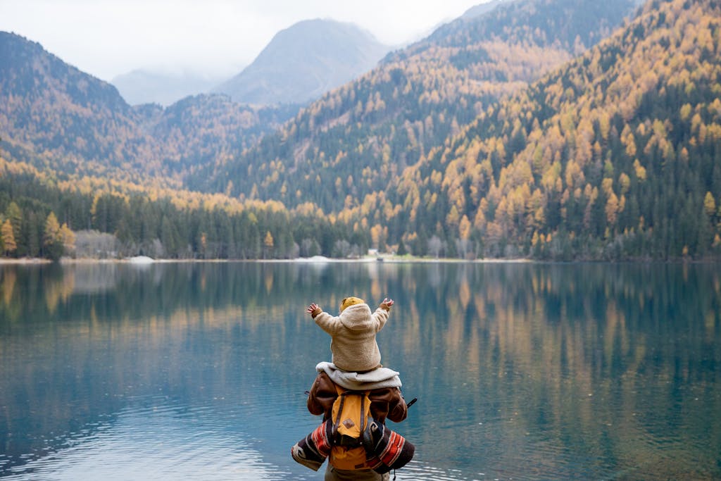 A parent and child seated on a lakeside log, enjoying the serene mountain view in autumn.