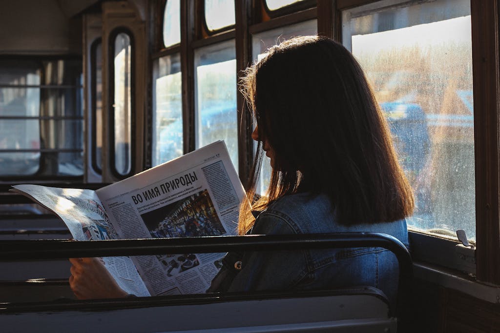 A woman reads a newspaper in a sunlit bus, capturing a moment of quiet reflection.