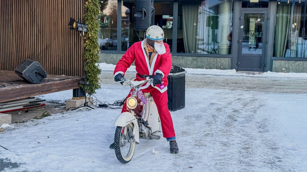 Santa Claus dressed in red riding a vintage scooter on snowy streets of Boden, Sweden.