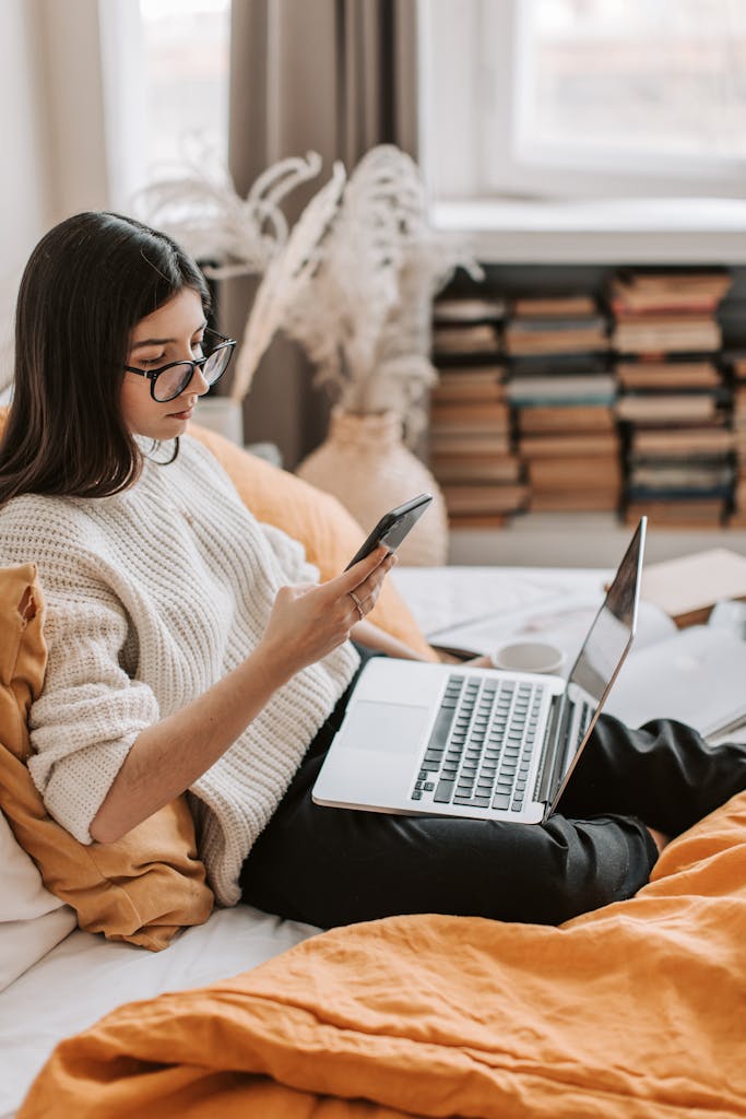 Side view of focused young female student sitting on bed at home while browsing Internet on laptop and using smartphone