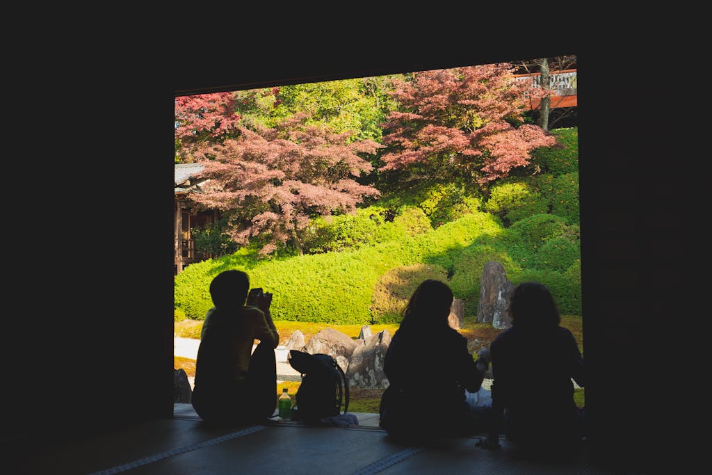 Silhouette of people admiring serene Japanese garden framed by temple window, showcasing lush greenery and colorful foliage.