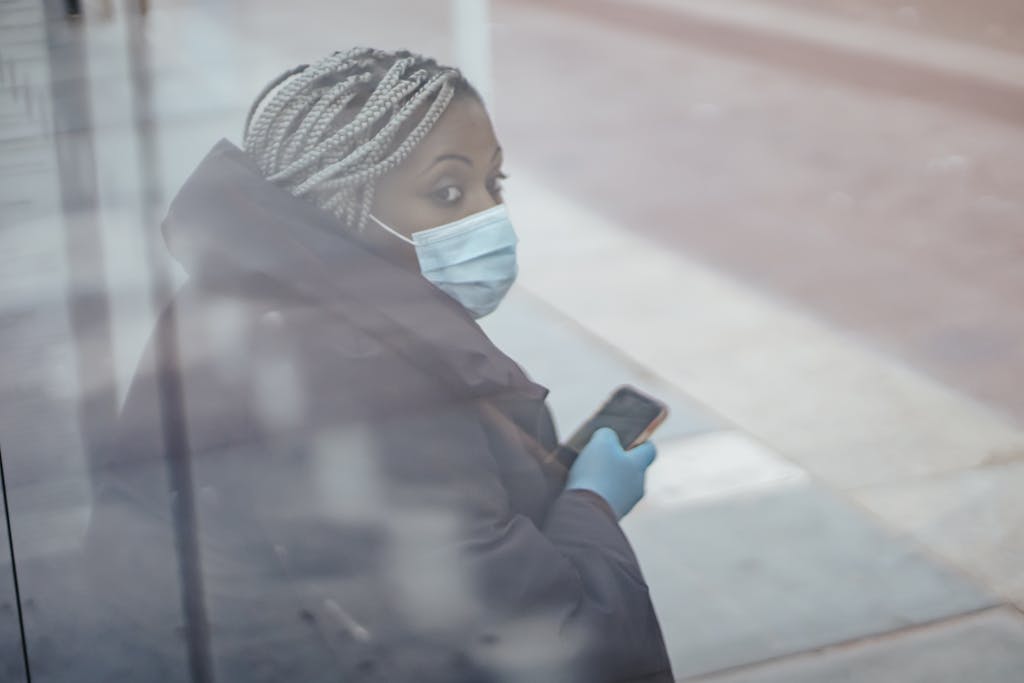 Through glass wall side view of mature ethnic female medic in outerwear with cellphone looking away in town