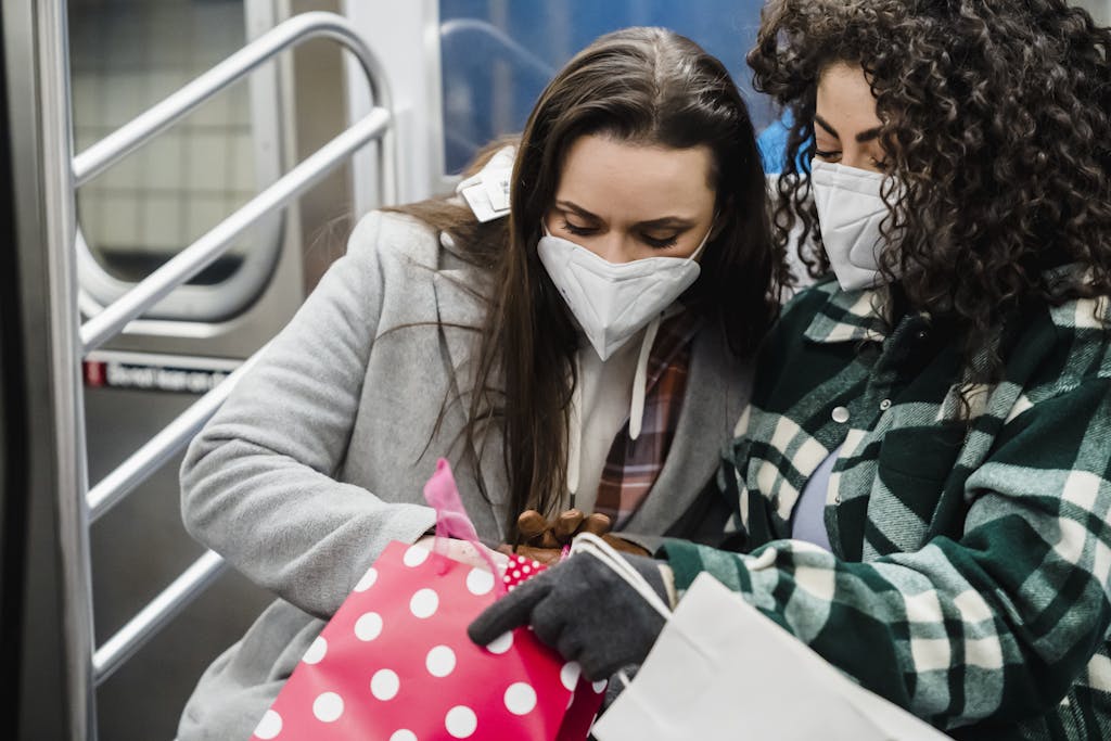Two women wearing masks on a subway seat, exploring shopping bags amidst pandemic precautions.