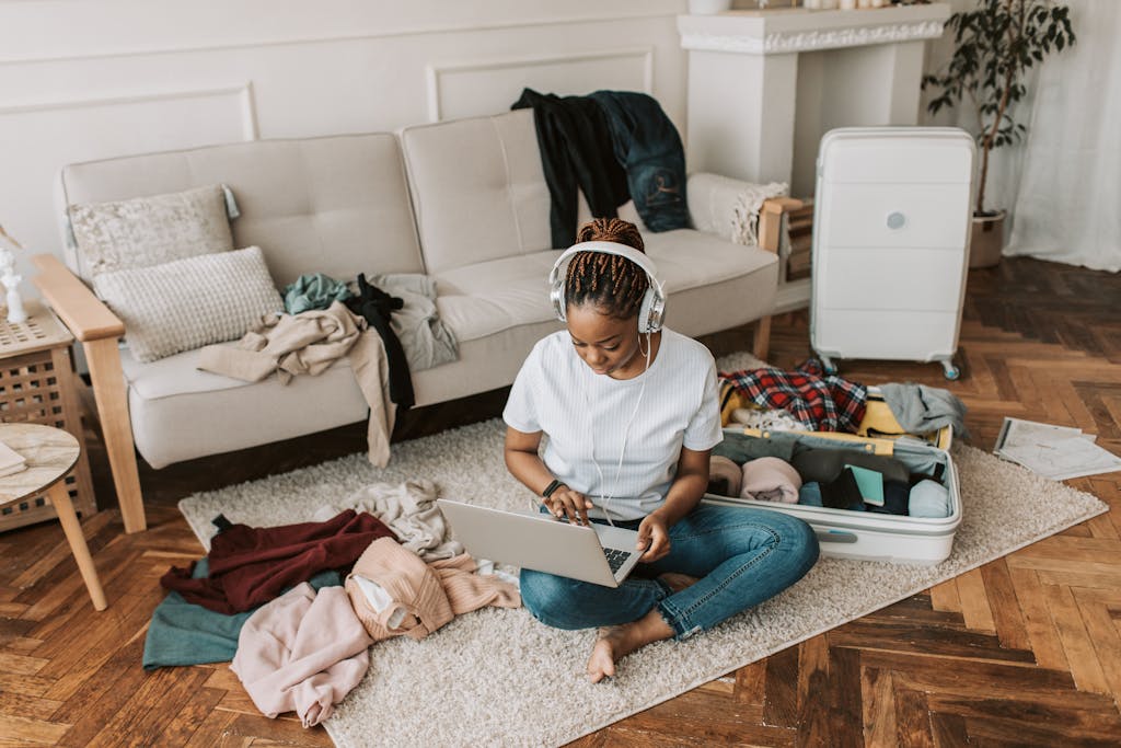 Young woman packing a suitcase in a living room while working on a laptop, preparing for travel.