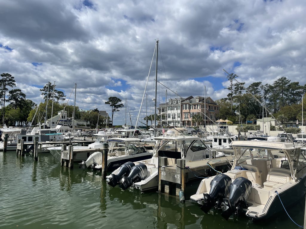Explore the tranquil marina at Hilton Head Island with docked sailboats and motorboats under a partly cloudy sky.